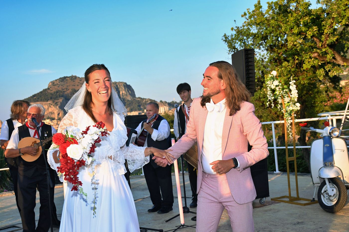 Due sposi ridono mentre il gruppo suona, con la Rocca di Cefalù alle spalle